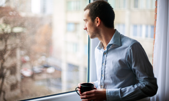 A man contemplating and looking out of his window while holding a cup of coffee.